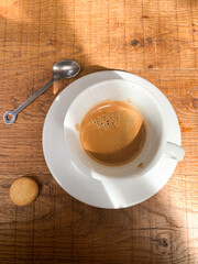 Top view of an espresso cup on a white saucer with a small biscuit and a teaspoon on a wooden table.