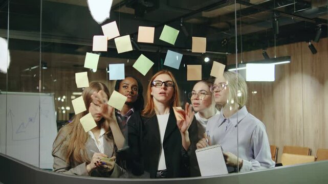 a diverse group of talented women collaborates in a modern office, using colorful sticky notes on a glass board to brainstorm and develop innovative ideas during focused discussions