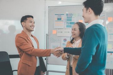 Two businessmen holding hands in conference room, team of analysts celebrating after achieving great success in business strategy.