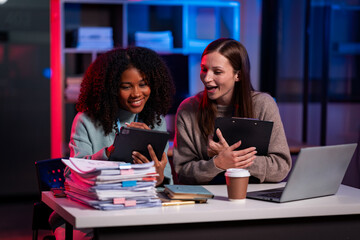 Two businesswomen working overtime, chatting in the office, working with laptops, documents, graphs, financial analysis with lights in the office