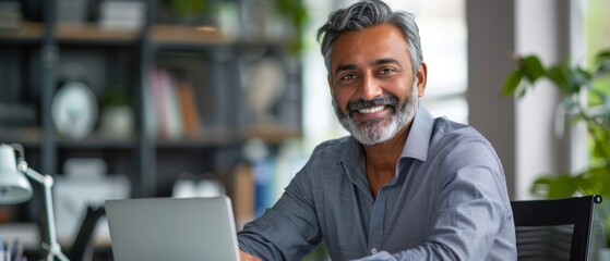 Smiling mature business man executive wearing shirt sitting at desk using laptop. Happy busy professional middle aged Indian businessman investor working on computer looking away in office. Copy space