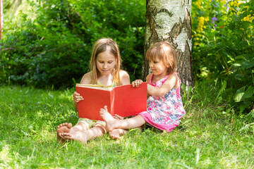 Two sisters reading a red book together in a sunny garden setting