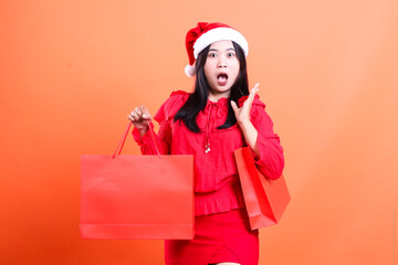 Charming woman wearing red dress and Santa hat, surprised to camera, hand carrying red bag of Christmas gifts and beside head, isolated on orange background