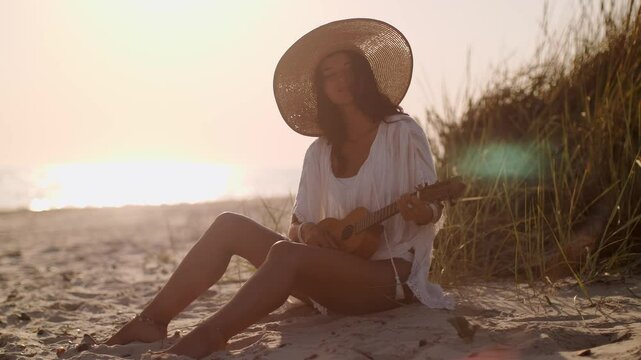 A carefree boho woman plays the ukulele on the beach at sunset, wearing a straw hat and white blouse. This tranquil scene embodies summer relaxation and artistic beach vibes.