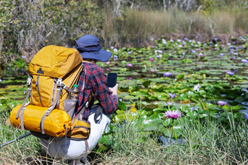 A hiker with a yellow backpack kneels by a tranquil water lily pond, capturing the beauty of nature...