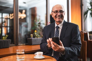 Smiling senior businessman with eyeglasses in elegant suit using mobile phone and drinking coffee while sitting in the cozy city cafe.