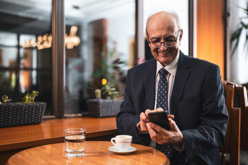 Happy senior businessman with eyeglasses in elegant suit using mobile phone and drinking coffee while sitting in the cozy city cafe.