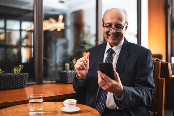 Happy and confident senior businessman with eyeglasses in elegant suit using mobile phone and drinking coffee while sitting in the cozy city cafe.