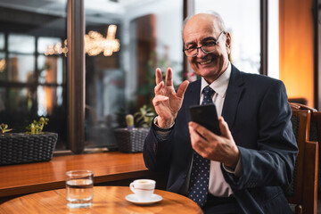 Smiling successful senior businessman with eyeglasses in elegant suit using mobile phone and drinking coffee while sitting in the cozy city cafe.
