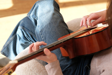 A young woman playing an acoustic guitar while seated comfortably in a cozy living room, surrounded by soft furnishings. Sunlight filters through the window, creating a warm atmosphere.