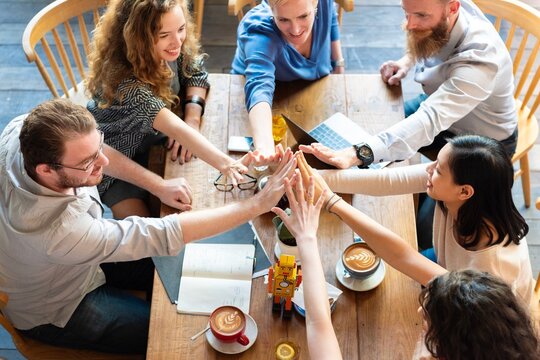 Group of diverse people high five around table with coffee and notebooks, showing teamwork and collaboration casual meeting setting. Group of diverse friends hands together over the table as teamwork