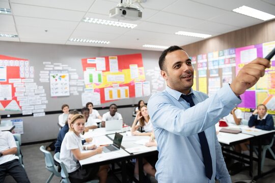 Classroom with diverse students and a male teacher pointing at board. Students engaged, teacher explaining. Classroom setting, teacher, students, learning. Teacher teaching a diverse high school class - Powered by Adobe