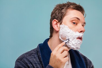 Young Caucasian man shaving, applying shaving cream, and using a razor. Shaving cream covers his...