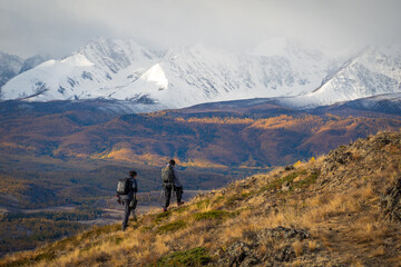 Two hikers walking in hilly terrain surrounded by autumn vegetation and snowy mountains under cloudy sky.