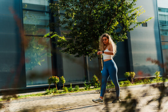 Young fit sportswoman with curly blonde hair wearing white top and blue leggings checking time on her smartwatch while walking on a sidewalk near a modern glass building on a sunny day