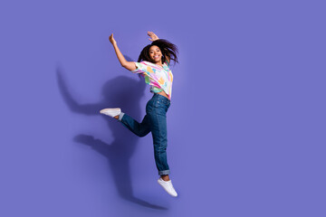 Young woman in colorful shirt and jeans joyfully jumping against a vibrant violet background,...