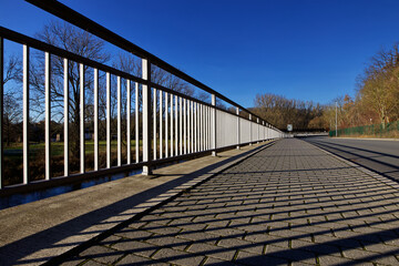 Obraz premium Brückengeländer im Sonnenlicht an Strasse mit Bäumen, Licht und Schatten, markante Form Design, blauer Himmel Blickwinkel von vorn, ruhige Szene in Europa Deutschland Thüringen Gera