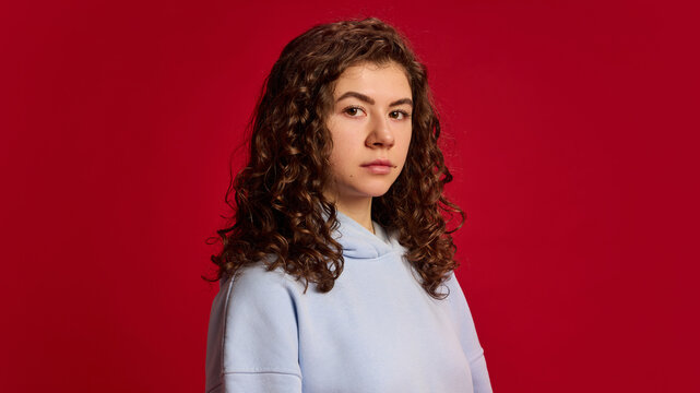 Close up of young attractive lady with brunette curly hair looking at camera with serious expression against vibrant red background. Concept of human emotions, beauty, fashion and style. - Powered by Adobe