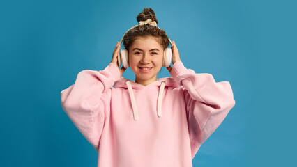Portrait of positive, smiling young woman dressed pink hoodie adjusting headphones and look at camera against blue studio background. Concept of human emotions, beauty, fashion and style.