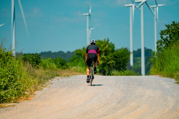Obraz premium Cyclist riding on gravel road surrounded by wind turbines under clear blue sky in summer afternoon