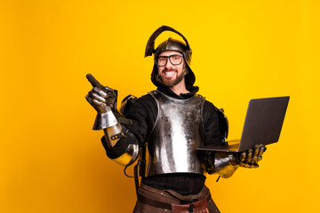 Young man wearing medieval knight armor holding a laptop with a cheerful expression against a...