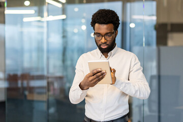 Serious thinking man standing with tablet computer in the middle of the office, businessman working in the evening, using online application to view online financial investment reports.