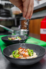 A close-up shot of a chef’s hand sprinkling cheese over a gourmet dish featuring salmon, broccoli, and grains, set in a modern kitchen environment.