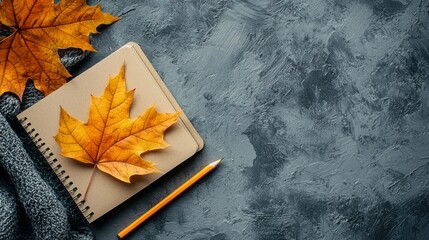 A delightful autumn composition showcases a vibrant golden leaf resting gracefully on an open notebook. A cozy texture from a knitted scarf complements the warm hues, inviting creativity