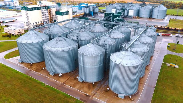 Approaching a group of metal elevators standing in four rows. Large territory of an agricultural plant functioning at backdrop. Aerial view.