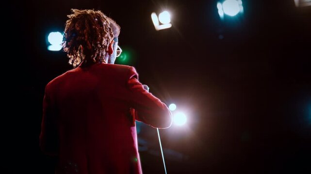 Stylish black man with dreadlocks performing on stage of concert hall, back view . Stand-up comedian or host of event, presentation of new business project on conference, back view of african man