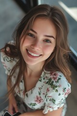 Obraz premium A young woman posing with a smile, wearing a flowery blouse and casually holding her phone in hand. Her hair is playfully flipped to one side.