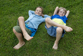 two cute barefoot boys 7 - 8 years old lie on their backs on the grass. Happy childhood, positive emotions, friendship, joy. Friendly children, brothers having fun, playing outdoors. Summer holidays