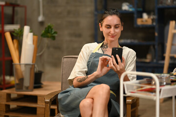 An artist in an apron sits in a cozy studio, smiling while using her smartphone, with art tools and supplies around her, taking a moment to connect digitally