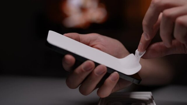 Male hands carefully peeling off protective film from screen of new smartphone, revealing pristine surface underneath in soft diffused lighting in dark room, close-up. Shooting in slow motion.