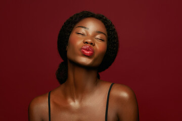 Carefree confident African American woman with natural curly hair blowing a kiss, wearing red lipstick and a black strap top against a bright background.
