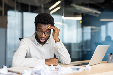 Disappointed and upset man doing paperwork inside office. Unsatisfied with financial reporting results sitting near crumpled pieces of papers. Businessman with laptop among documents.
