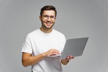 Young man with glasses smiles while using a laptop in a minimalist indoor setting