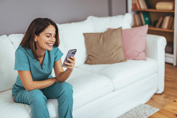 Smiling Woman in Scrubs Using Smartphone on Comfortable White Sofa