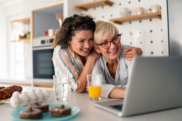 A joyful mid-age Caucasian daughter and her senior mother in a modern kitchen share a heartwarming...