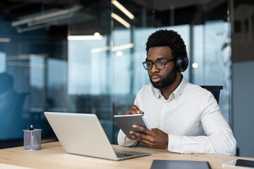 Obraz premium Man watching online video meeting conference, business meeting online. Worker with laptop and headphones writing data in notebook sitting at desk inside office.