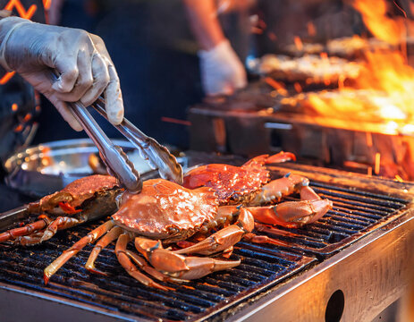 Crabs being grilled over an open flame, with tongs and gloved hands handling them, creating a lively barbecue scene.