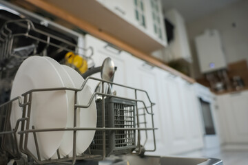 Open dishwasher close-up with clean white plates and cutlery on the left of the photo against the background of a white wooden kitchen out of focus. The concept of washing dishes, dishwashers