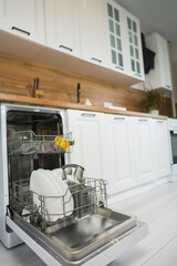 Vertical photo of open dishwasher department with clean plates and cutlery against light kitchen background with white cabinets and wooden countertop and panel. The concept of washing dishes