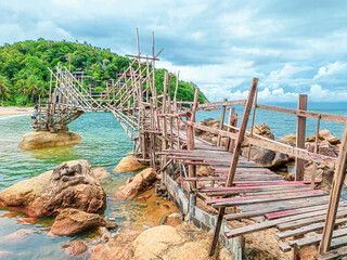 Obraz premium Rustic Coastal Fishing Pier: A weathered wooden pier extends over tranquil turquoise waters, leading towards a lush green island under a dramatic sky.