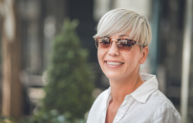 Closeup face of european blond adult woman wearing sunglasses. Smiling face of beautiful mid aged female person dressed in white summer shirt. Outdoors