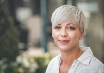 Close-up portrait of european happy blond adult woman with short blond hair. Face of beautiful female person dressed in white summer shirt. Outdoors