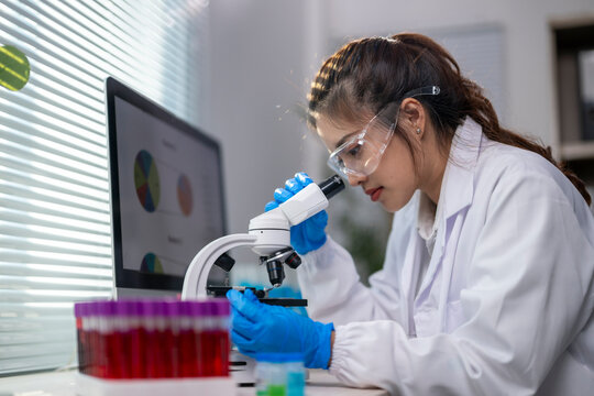 A woman wearing a lab coat and goggles is looking through a microscope