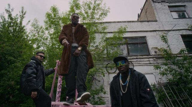 Low angle full shot of Black male rapper in brown fur coat and pants vibing to hip-hop music with his diverse friends in black baggy clothes and looking at camera, filming music video outdoors