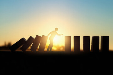 Backlit business man stopping abstract wooden domino blocks on bright sunset sky background with mock up place. Crisis, recession and challenge concept. 