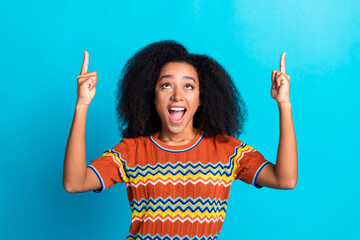 Joyful young woman with curly hair pointing upwards against a vibrant blue background in stylish...